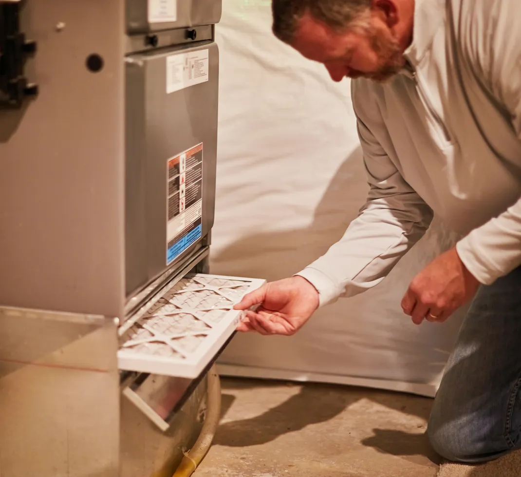 A person replacing a furnace air filter in an HVAC system, kneeling in front of the unit to insert a new filter into the filter slot.