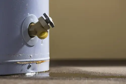 A low-angle close-up view of water leaking from the plastic faucet on a residential electric water heater sitting on a concrete floor in the basement.
