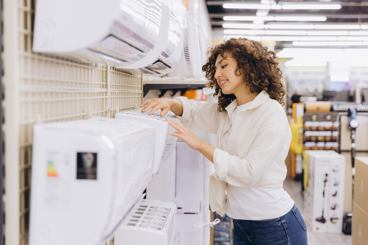 Woman comparing wall-mounted air conditioning units in an appliance store while choosing the best AC system for home cooling needs.