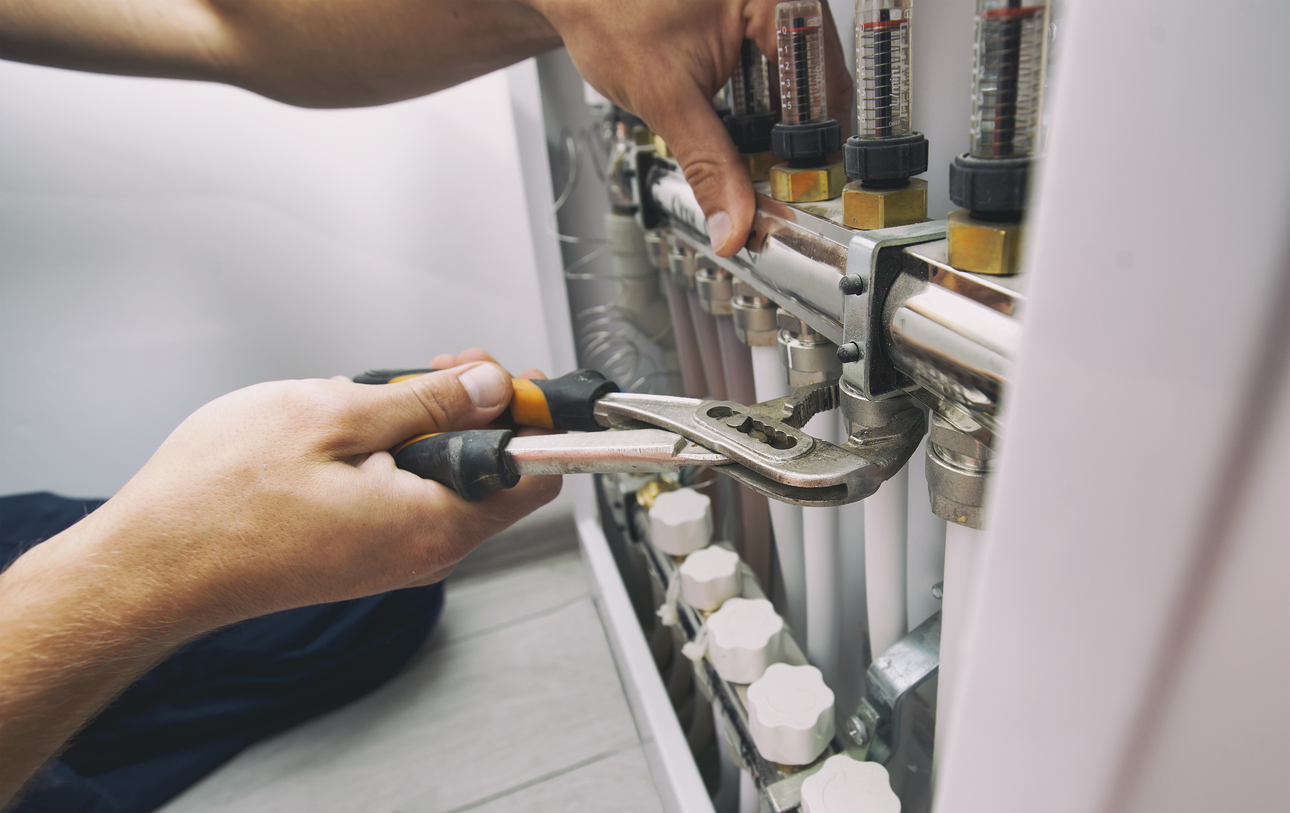 Close-up of a technician’s hands using pliers to tighten a fitting on a heating system manifold with flow meters and valve controls.
